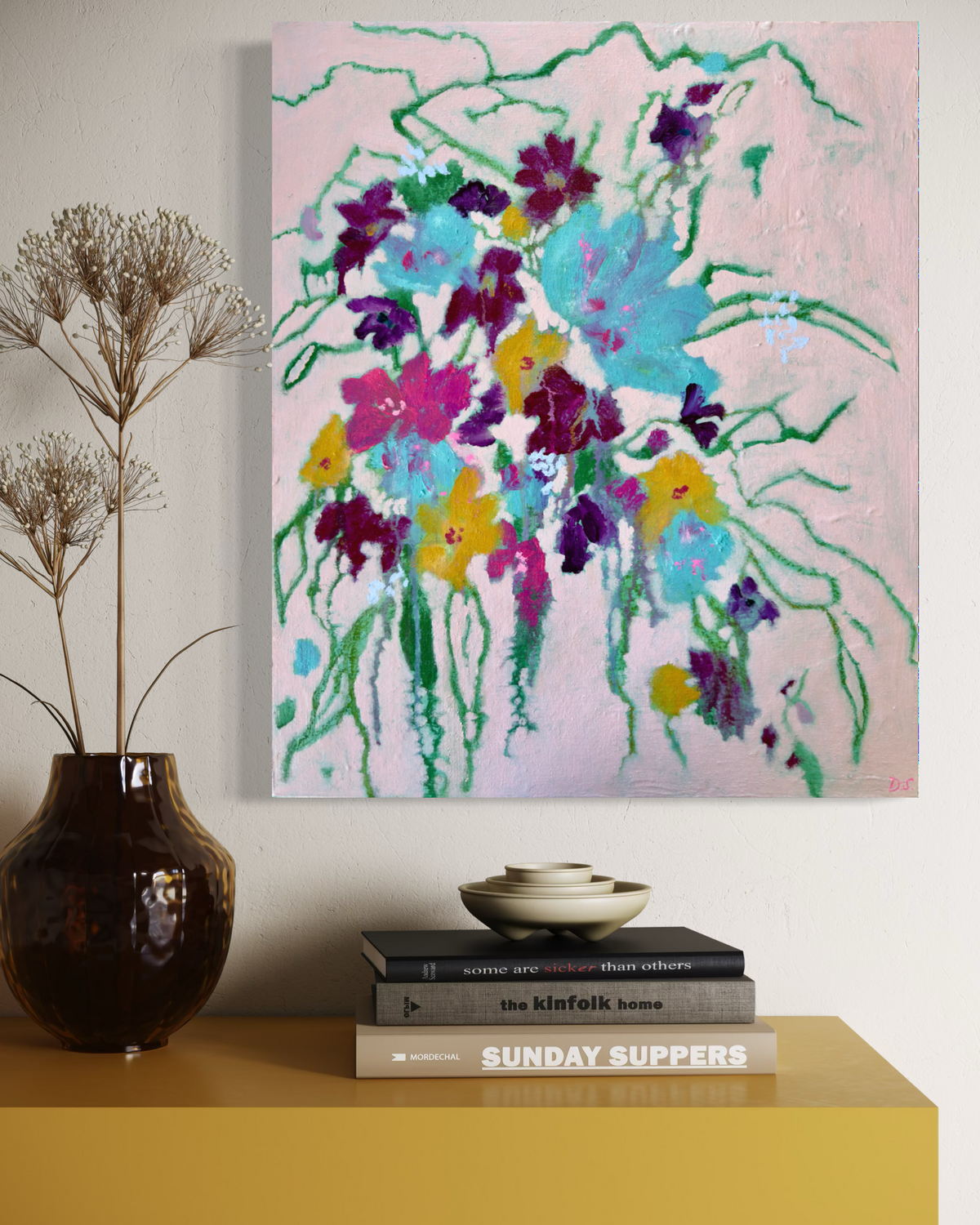 A colorful abstract floral canvas painting hanging on a light beige wall above a yellow sideboard, styled with a dark brown vase of dried grass and a stack of books.