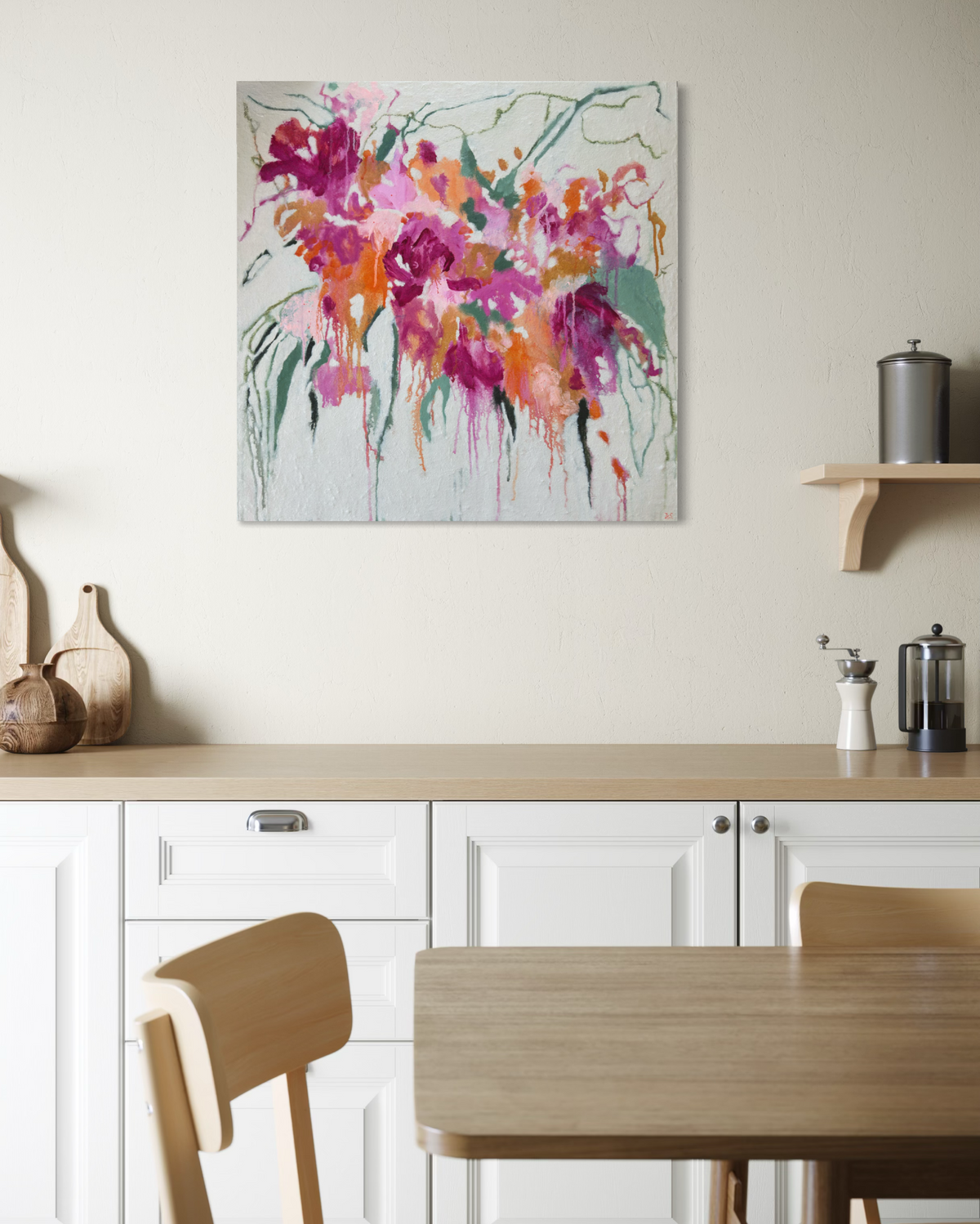 An abstract floral canvas print with vibrant magenta, pink, orange, and green brushstrokes on a light background, displayed on a beige wall above a modern white kitchen counter with a wooden table and chairs.