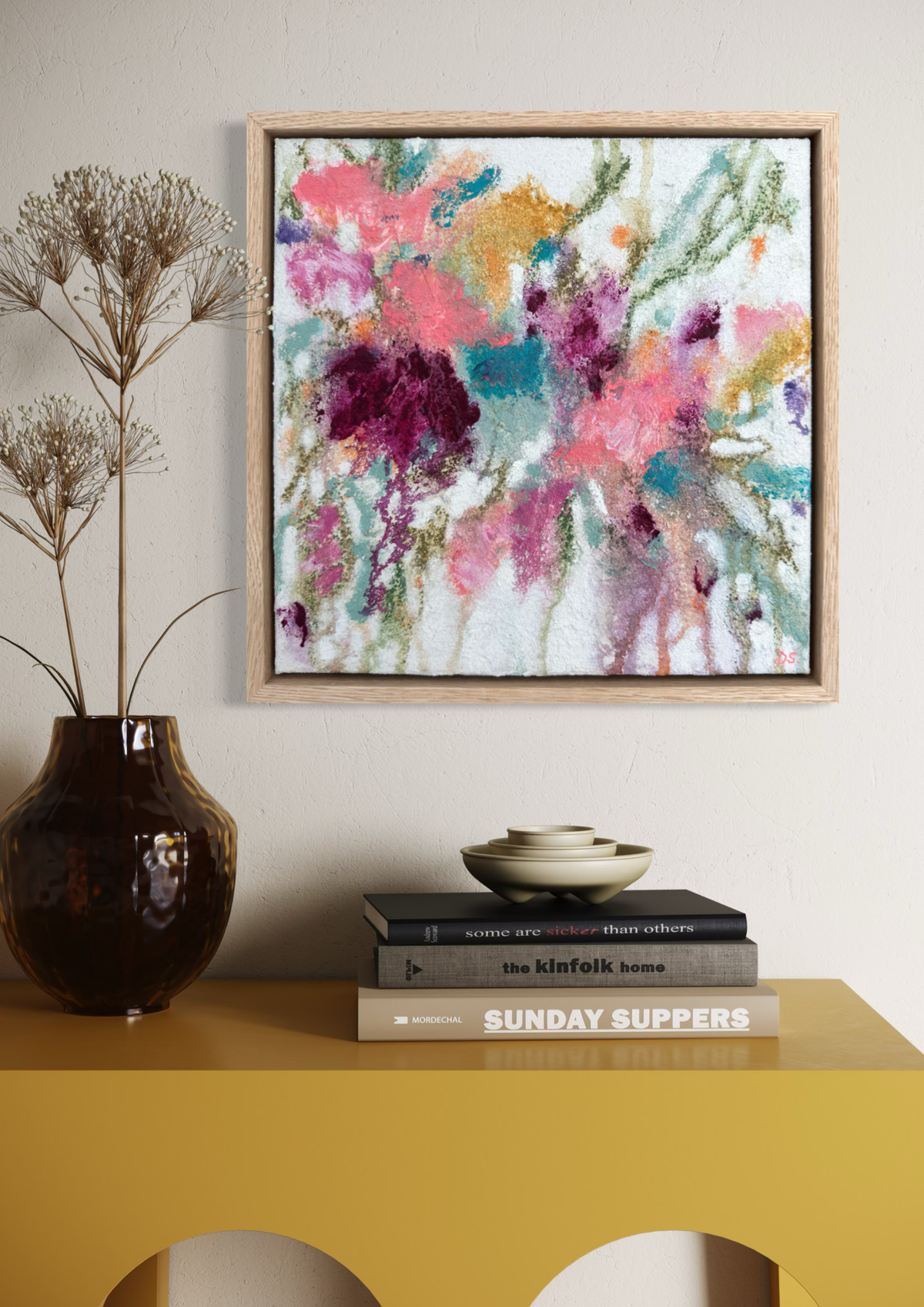 A framed abstract floral painting with pink, purple, teal, and orange brushstrokes hangs on a light beige wall above a mustard yellow console table. The table holds a dark brown vase with dried stems, a stack of books, and a small bowl.
